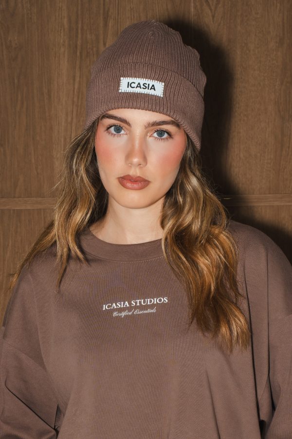 Female model wearing a mocha chocolate colour beanie knit beanie. The beanie has a sewn in brand logo label which is black and white.